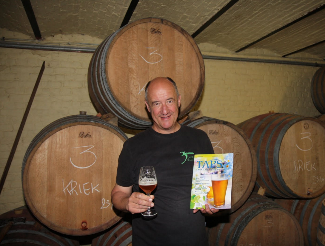 Armand Debelder with a glass of lambic and barrels filled with kriek behind him. 